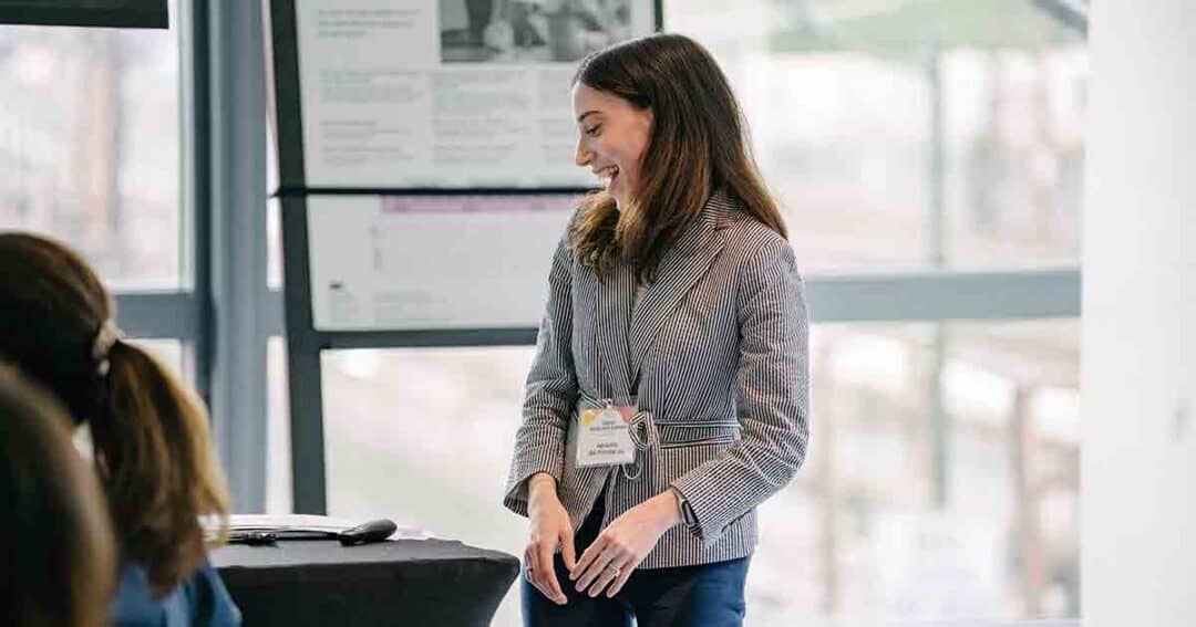 A woman in a striped blazer with a conference badge smiles while standing at the front of a room, appearing to give a presentation. There are people seated nearby, and posters are displayed in the background.