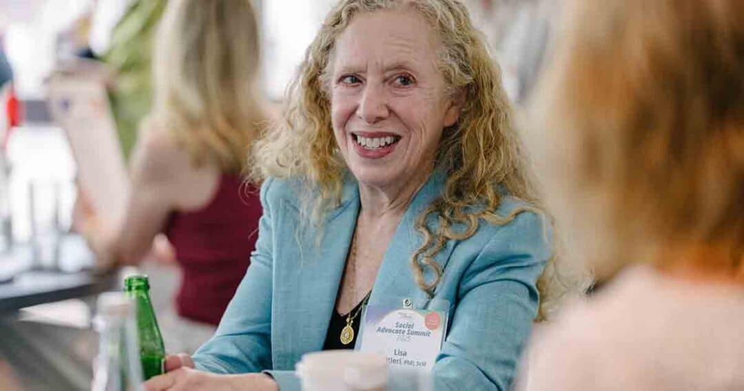 A woman with curly blonde hair wearing a blue blazer smiles while sitting at a table. She has a name tag and holds a green bottle. Other people are visible in the blurred background.