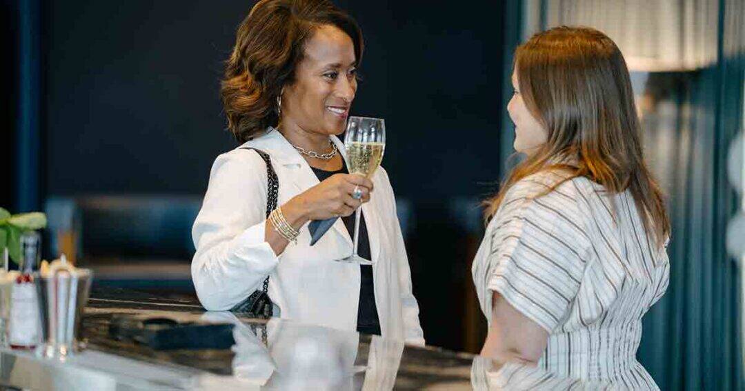 Two women stand at a bar, conversing. One woman in a white blazer smiles and holds a glass of champagne, while the other, wearing a striped top, listens attentively. The setting appears relaxed and upscale.