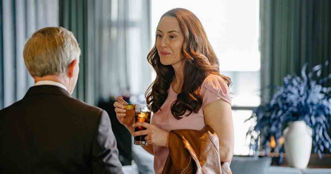 A woman holding a drink and a brown jacket talks to a man in a suit inside a room with large windows and blue curtains. A leafy plant is visible in the background.