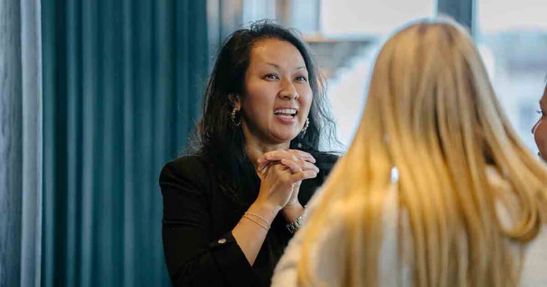 A woman with dark hair smiles and clasps her hands while talking to two blonde women in a brightly lit indoor setting.