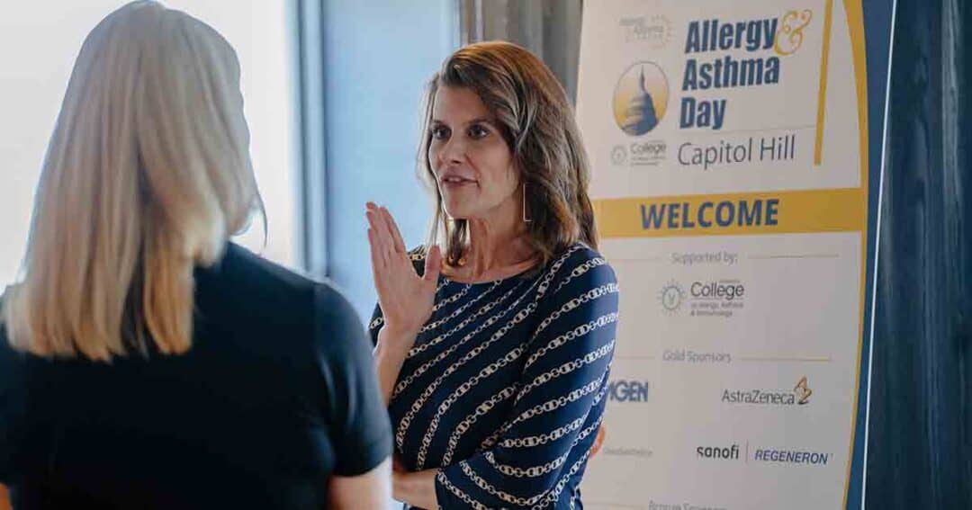 Two women talk in front of a sign that reads “Allergy & Asthma Day on Capitol Hill” with sponsor logos below. One woman gestures with her hand while speaking.