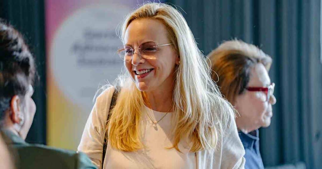 A woman with long blonde hair and glasses smiles while talking to others indoors. Two other people are partially visible in the background. The setting appears casual and friendly.