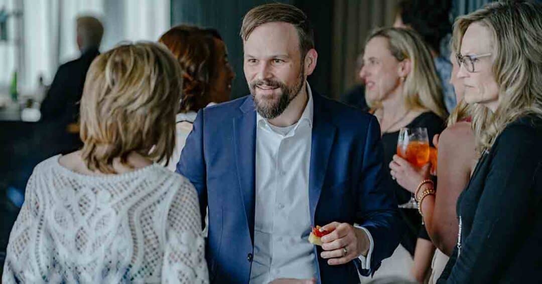 A man in a blue suit smiles and talks with a group of women at a social gathering; he is holding a small appetizer, and people around him are holding drinks.