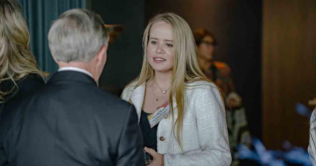 A young woman with long blonde hair, wearing a white jacket and name tag, is speaking to an older man in a suit at an indoor event. Other people are blurred in the background.