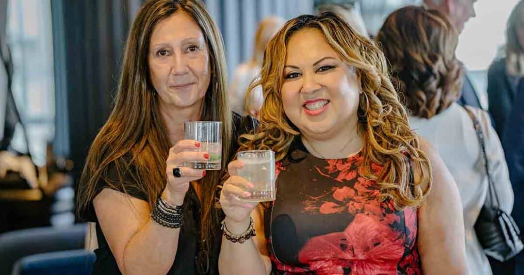 Two women smiling and holding drinks, posing together at an indoor social gathering with other people in the background.