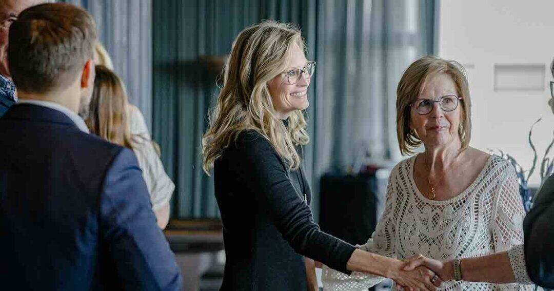 Two women smiling and shaking hands at an indoor gathering, surrounded by other people in business or semi-formal attire. The setting appears to be a professional or social event.