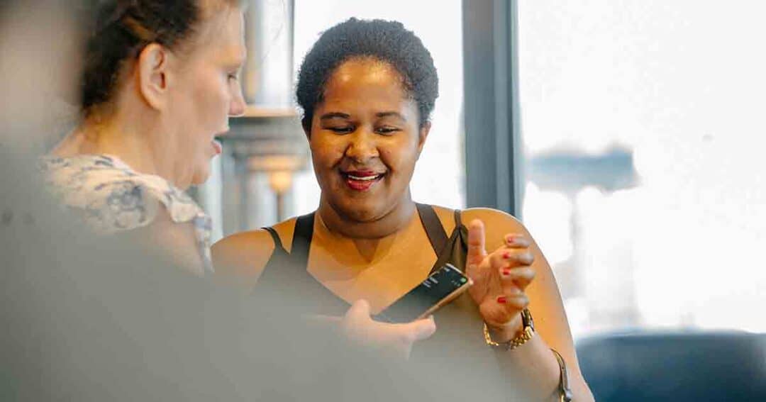 Two women smiling and looking at a smartphone together indoors, one holding the phone while the other gestures, with natural light coming through the window behind them.