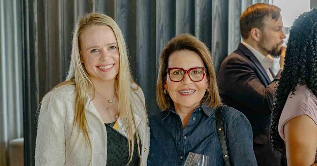 Two women smile at the camera at a social event, one with long blonde hair and the other with short brown hair and red glasses. People are mingling in the background.