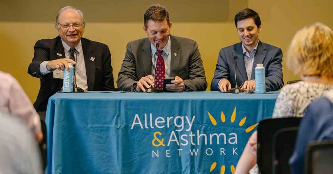 Three men in suits sit behind a table with an "Allergy & Asthma Network" tablecloth, speaking and smiling at an event, facing an audience. Bottles of water and microphones are on the table.