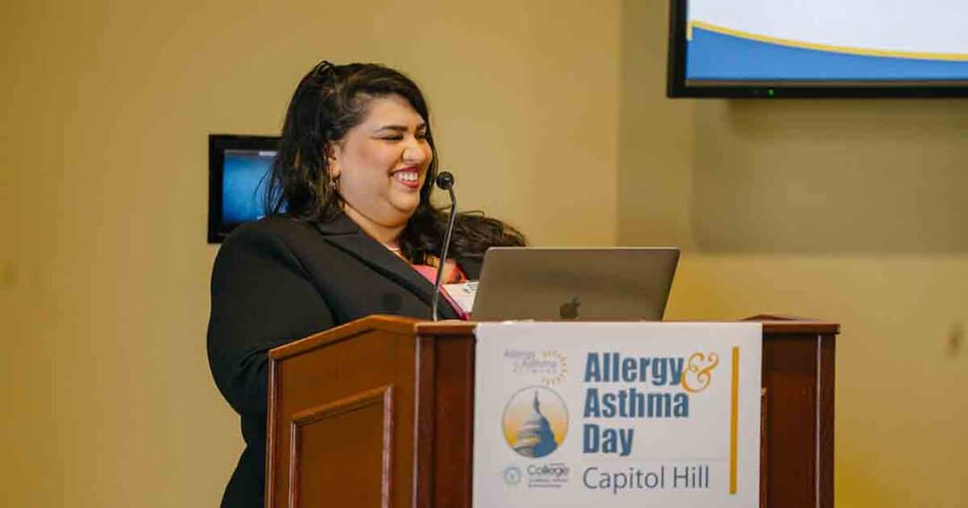 A woman smiling stands at a podium with a laptop, speaking at an event labeled "Allergy & Asthma Day Capitol Hill." A presentation screen is partially visible in the background.