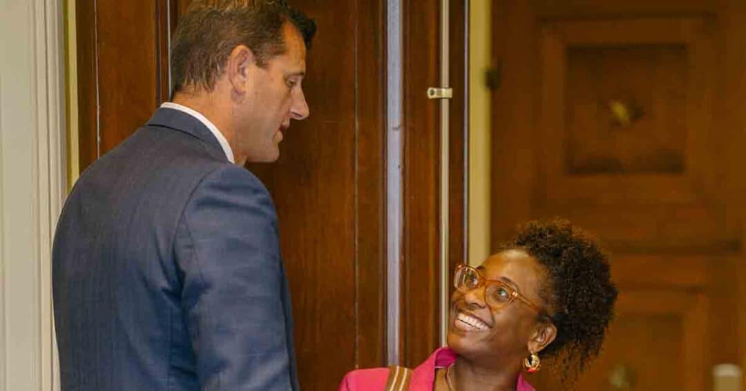 A man in a blue suit stands by a doorway, talking with a smiling woman wearing glasses and a pink blazer. The woman looks up at him, and they appear to be having a friendly conversation indoors.