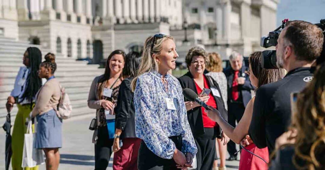A woman is being interviewed with a microphone by a reporter in front of a government building, with a group of people standing and talking in the background on the steps.