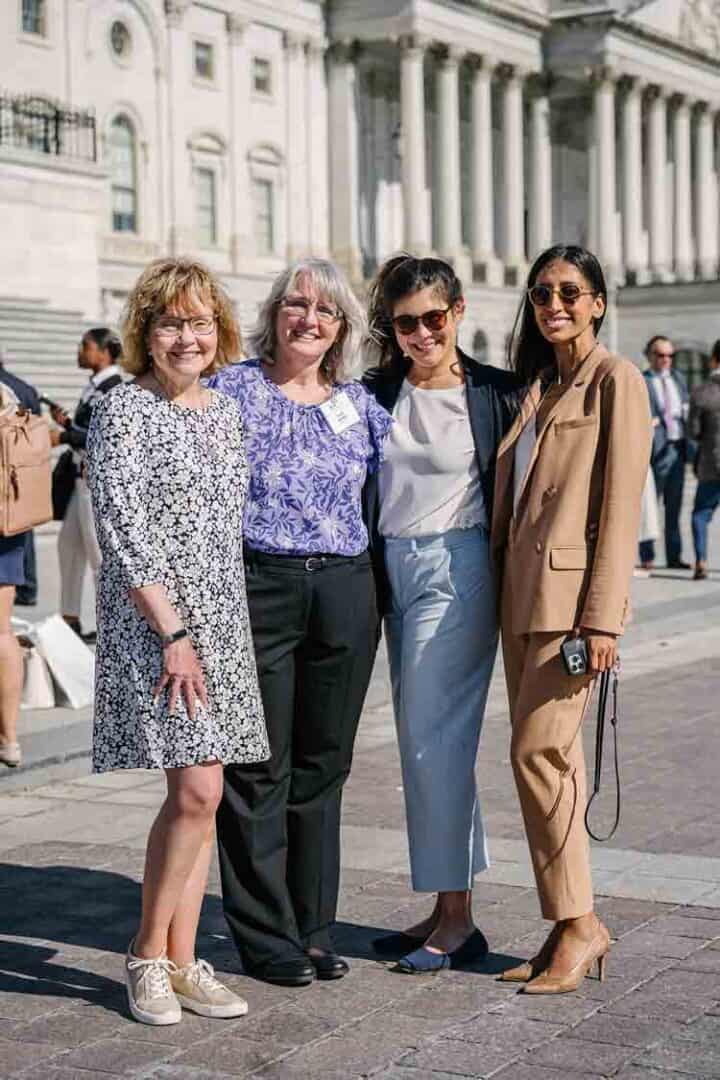 Four women stand together smiling in front of a grand, white government building with columns. They are dressed in business casual and professional attire, and the scene is outdoors on a sunny day.