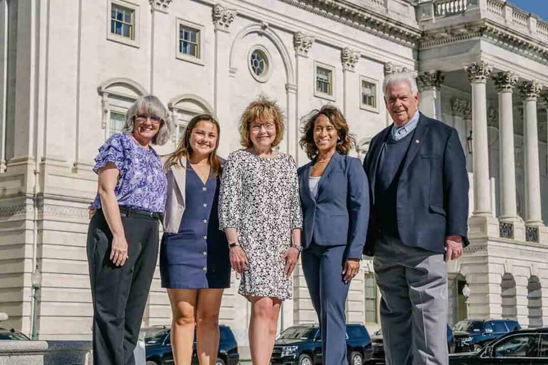 Five adults stand smiling in business attire in front of a large, white government building with columns and arched windows on a sunny day.