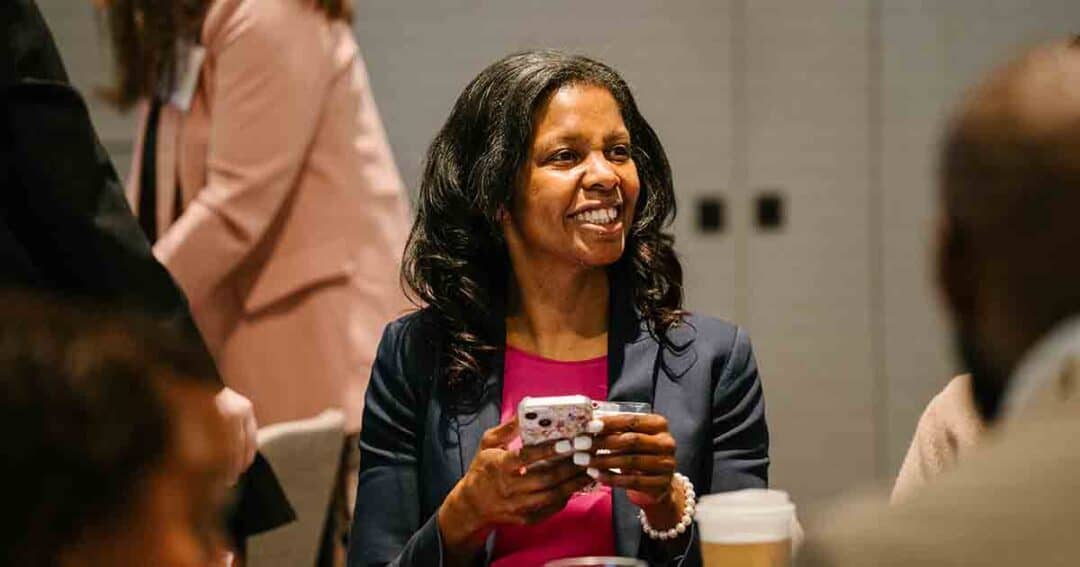 A woman in a navy blazer and pink top smiles while holding a smartphone, seated at a table with a coffee cup. Other people are visible in the background, suggesting a social or professional event.