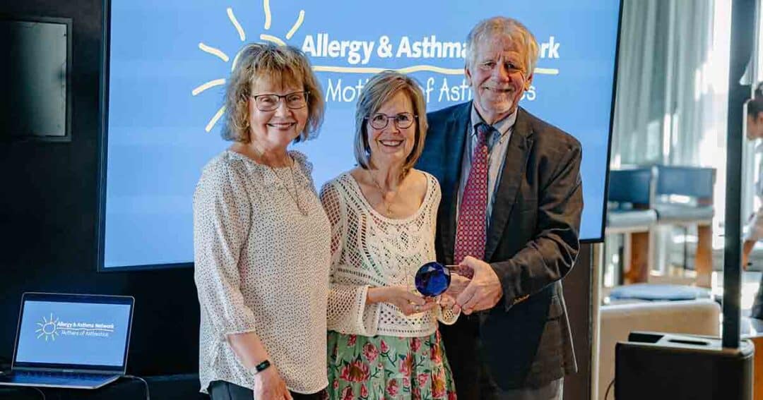 Three smiling adults stand together indoors; one woman in the center holds a blue glass award. A screen behind them displays "Allergy & Asthma Network." A laptop with the same logo sits on a table nearby.