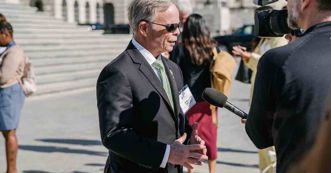 A man in a suit and sunglasses is being interviewed outdoors by a person holding a microphone and a video camera, with several people and steps visible in the background.