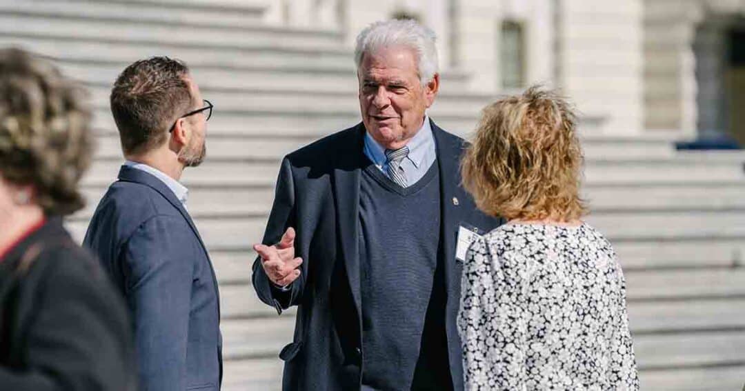 Three people stand outdoors on stone steps, engaged in conversation. The man in the center gestures while speaking, wearing a suit and sweater; the others listen attentively. The background is out of focus.