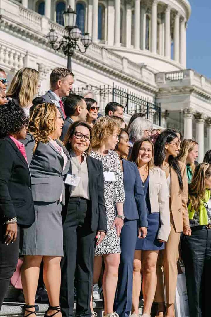 A diverse group of professionally dressed people stand together on the steps outside a large government building with columns, smiling and looking toward the camera.