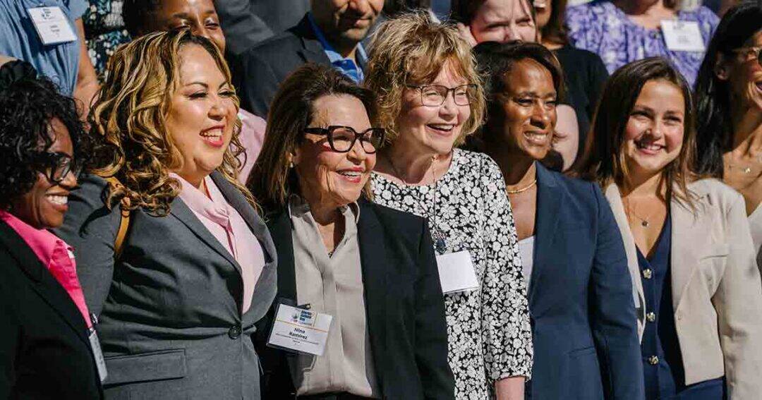 A diverse group of professionally dressed women stand closely together, smiling outdoors at what appears to be a conference or business event, with name badges visible on some of them.