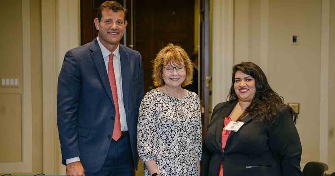 Three people pose indoors, dressed in business attire. The man on the left wears a suit and red tie, the woman in the middle wears glasses and a floral blouse, and the woman on the right wears a black blazer and red top.