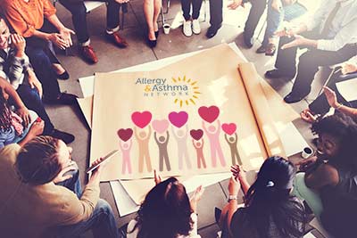 A diverse group of people sit in a circle, discussing or collaborating. In the center, there's a large poster with the Allergy & Asthma Network logo and colorful figures holding heart shapes.