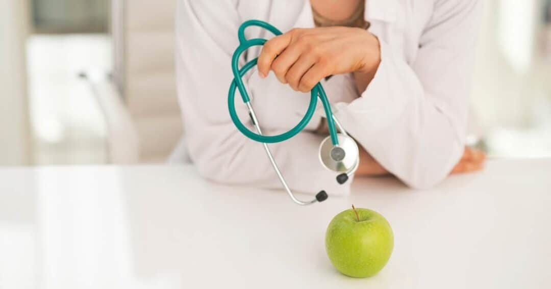 A person in a white coat holding a stethoscope is leaning on a table. In front of them, there is a green apple on the table.