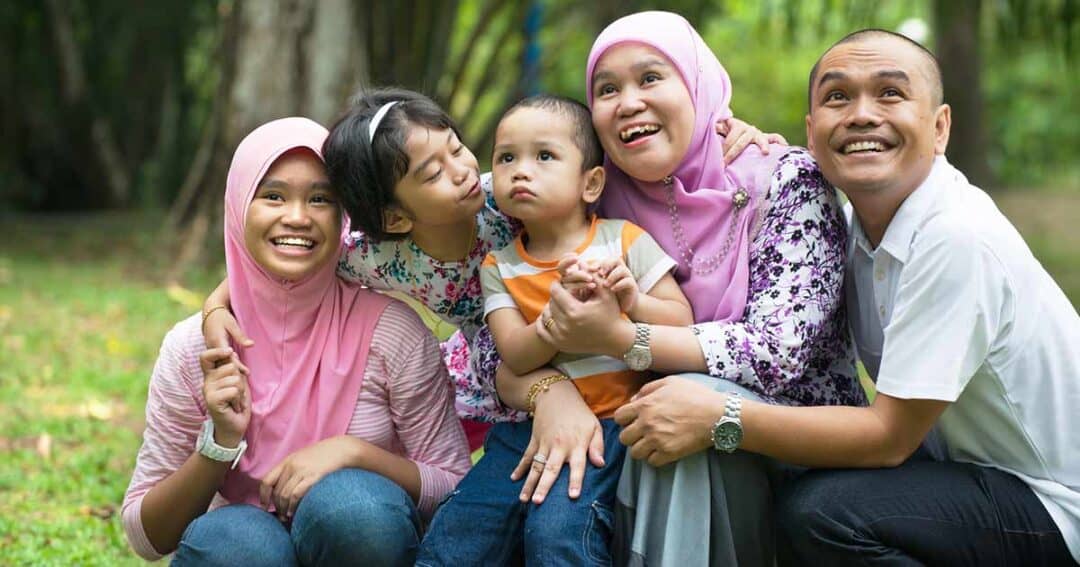 Health Equity in Allergies, Asthma and Immunology 10 A joyful family sits together on grass in a park. Two women in pink headscarves, a girl kissing a toddler’s cheek, and a smiling man all pose closely, exhibiting a sense of togetherness and happiness amidst lush greenery.