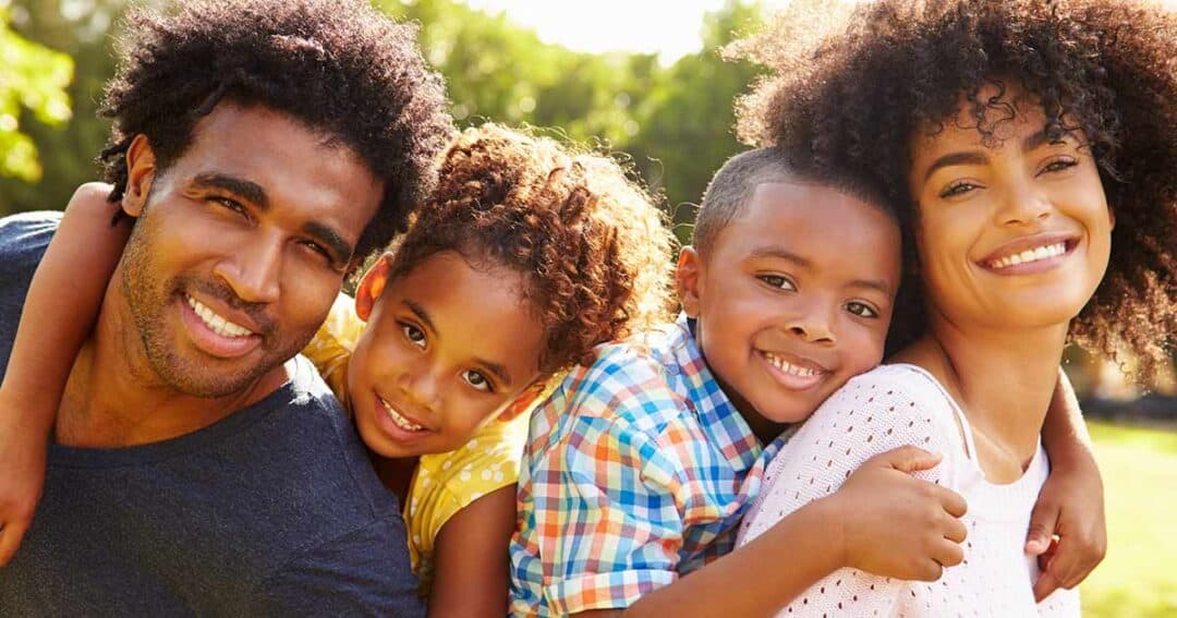 Health Equity in Allergies, Asthma and Immunology 7 A happy family of four smiling outdoors. The parents are embracing their two children, a daughter and a son. They are all wearing casual clothing and appear content, with greenery visible in the background.