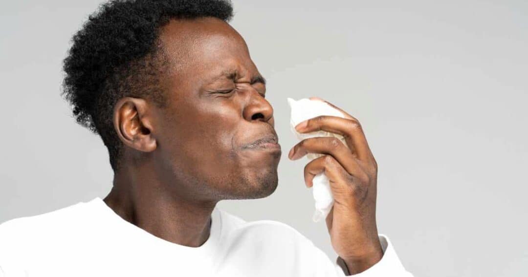 A person sneezing into a tissue, eyes closed, wearing a white shirt, against a gray background.