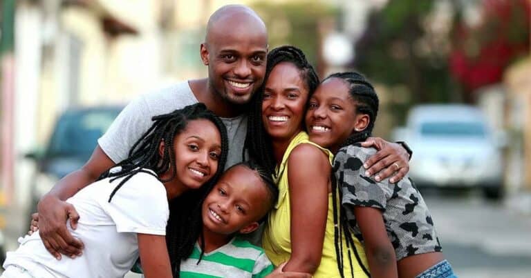 A happy family of five, including two adults and three children, stands closely together outdoors. They are all smiling, with warm expressions, in a casual setting with blurred buildings in the background.