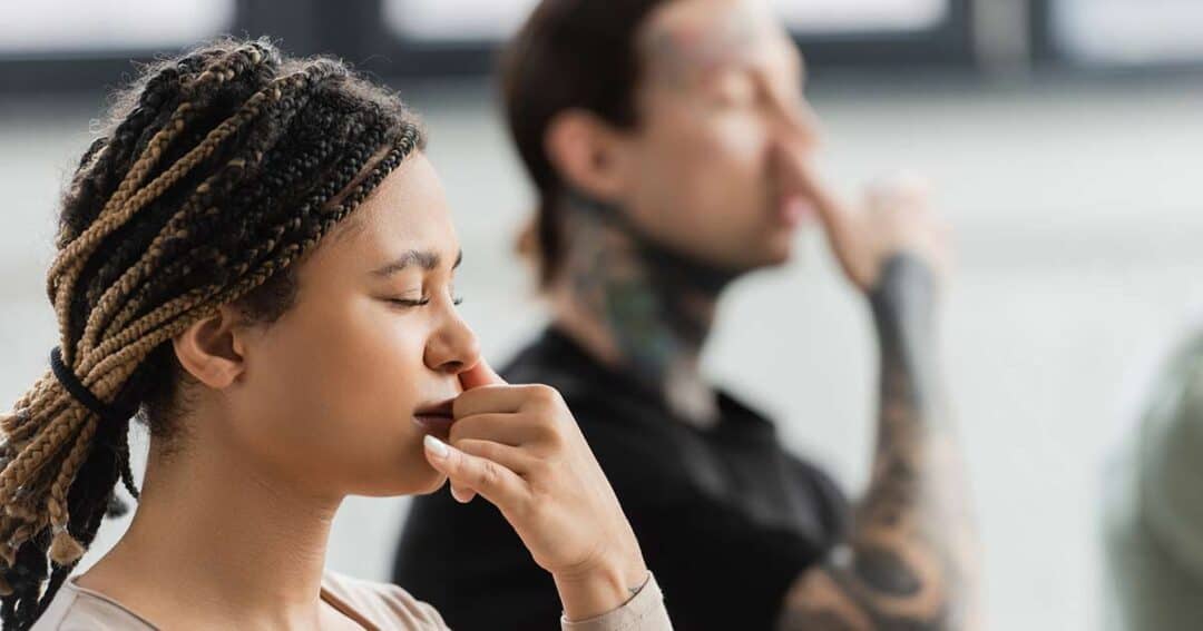 Two people are practicing alternate nostril breathing. The person in the foreground has braided hair and appears focused, while the person in the background has tattoos and is also engaged in the breathing exercise.