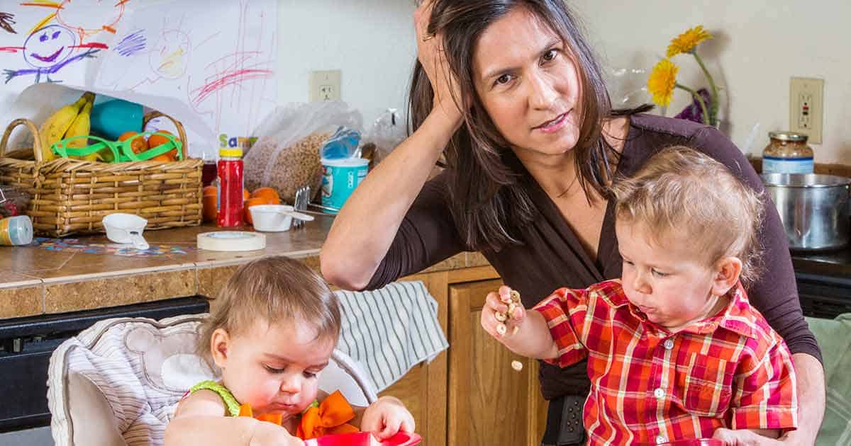 Mental Health and Living with Food Allergies 5 A woman sits at a kitchen counter with her hand on her head, looking tired. She is next to two toddlers, one playing with a red toy and the other holding food. The background includes colorful drawings and kitchen items.