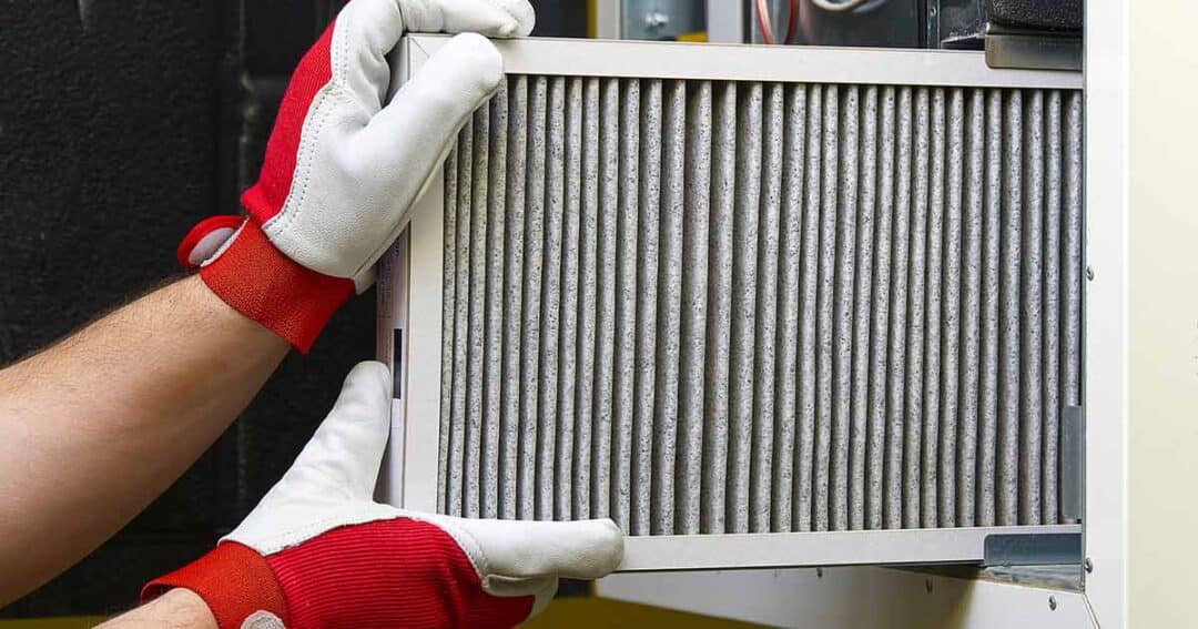 A person wearing red and white gloves is installing or removing an air filter from a ventilation system. The filter appears to be slightly dirty, indicating use. The background shows parts of the ventilation unit.