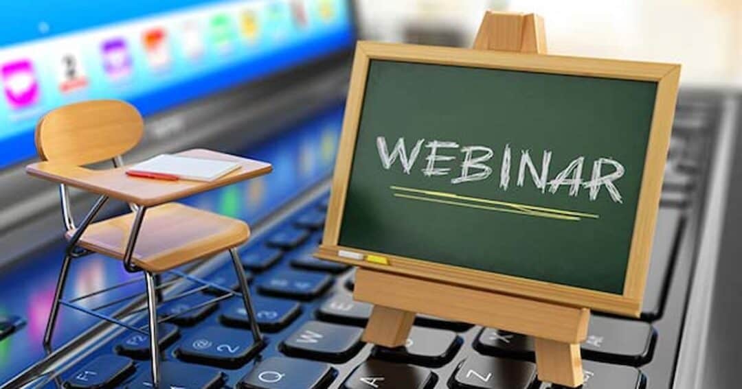 A miniature wooden desk and chair are placed on a laptop keyboard, adjacent to a small blackboard displaying the word "WEBINAR." The laptop screen shows colorful icons, suggesting an online learning or virtual seminar theme.