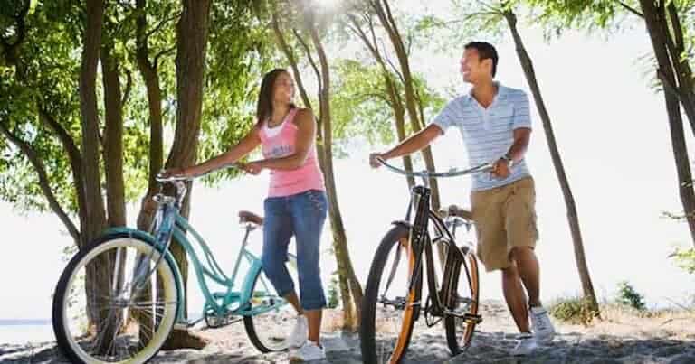 A woman and a man walking with bicycles through a sunlit, wooded area. The woman, in a pink tank top, and the man, in a striped shirt, are smiling at each other. Sunlight filters through the trees, casting dappled shadows on the ground.