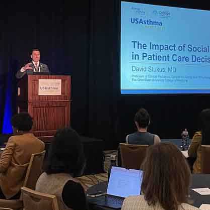USAsthma Summit 2023: Is Asthma Remission Possible? 4 A speaker at a conference stands at a podium delivering a presentation titled "The Impact of Social Media in Patient Care Decisions." The audience is seated, some with laptops open, in a dimly lit room.