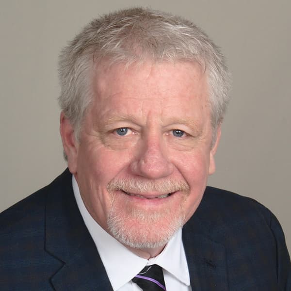 A smiling man with gray hair and a beard, wearing a dark suit and a light-colored shirt, appears against a plain background. He has a friendly expression and is positioned facing the camera.