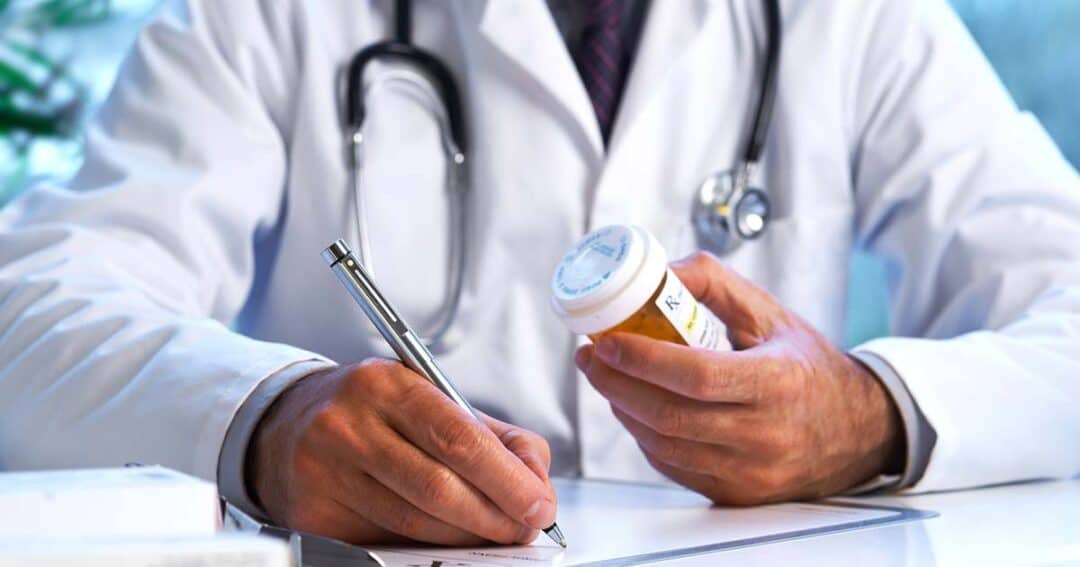 A doctor in a white coat writes a prescription while holding a pill bottle, with a stethoscope draped around their neck. Only the doctor's hands and partial torso are visible.