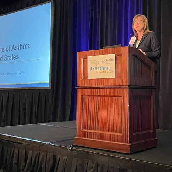 USAsthma Summit 2023: Is Asthma Remission Possible? 3 A woman stands at a podium with a microphone, presenting at the USAsthma Summit 2023. A large screen next to her displays a slide titled "The State of Asthma in the United States." The setting is a conference room with dark curtains.