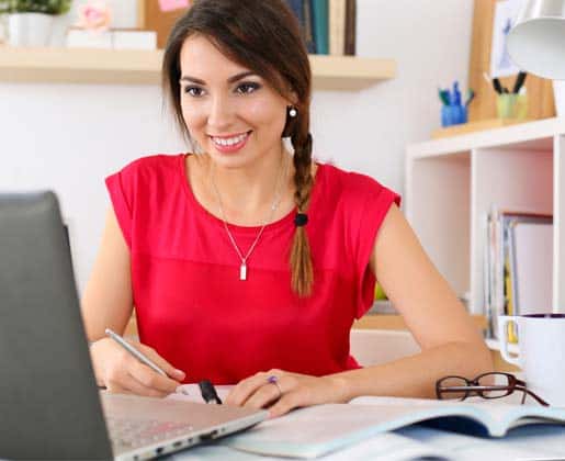 A woman with a braid and a red top sits at a desk, smiling while writing in a notebook. A laptop is open in front of her, with books, glasses, and a coffee mug nearby. The background has shelves with various office supplies.