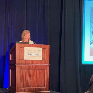 USAsthma Summit 2023: Is Asthma Remission Possible? 5 A person stands at a wooden podium with a sign that reads "US Asthma Summit 2023." They are speaking into a microphone on a stage with a dark curtain and a screen displaying a partial image on the right.