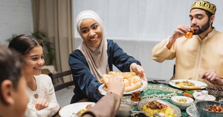 A family enjoys a meal together. A woman in a hijab smiles while passing a plate of food. A man in traditional attire drinks tea. A young girl is beside them, and the table is filled with various dishes.