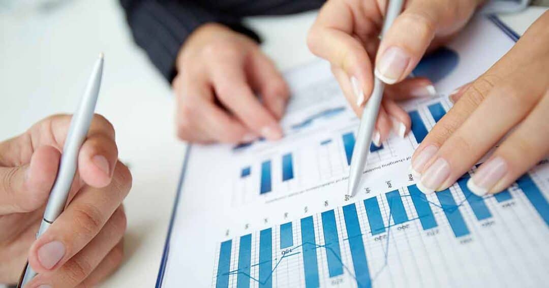 Financials and Accountability 1 Close-up of two people discussing a financial chart. One person holds a pen pointing at a bar graph with blue bars, while the other holds a pen nearby. The chart includes various data points and lines on a clipboard.