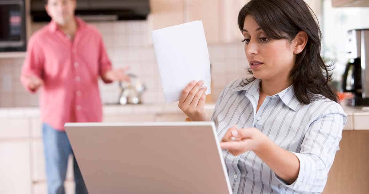 Mental Health and Living with Food Allergies 8 A woman sits at a table with a laptop, holding a piece of paper and looking concerned, perhaps considering the implications of food allergies. In the background, a man in a pink shirt stands with arms outstretched, appearing to speak about mental health support. They are in a kitchen setting.