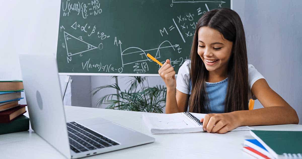 Mental Health and Living with Food Allergies 9 A girl is sitting at a desk with a laptop, notebooks, and her food allergy journal, focusing on learning food allergy tips. She is holding a pencil and smiling. In the background, a chalkboard brims with mathematical formulas and diagrams.