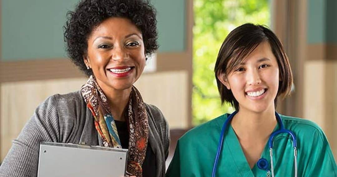 Health Equity in Allergies, Asthma and Immunology 11 Two smiling women are indoors. One wears business attire and holds a folder; the other is in green medical scrubs with a stethoscope around her neck. They stand in a bright room with natural light streaming in.