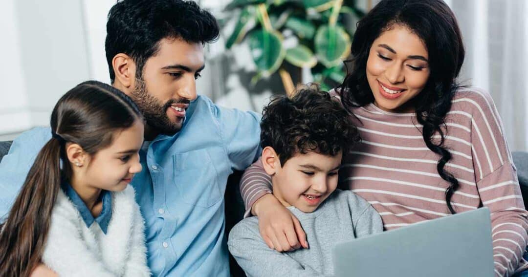 Health Equity in Allergies, Asthma and Immunology 8 A family of four sits together on a couch, smiling and looking at a laptop. The man and woman sit on either side of two children, a girl and a boy. A large green plant is visible in the background.
