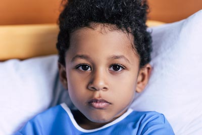 A young child with curly hair wearing a blue shirt looks directly at the camera while lying on a bed with white pillows and a wooden headboard. The expression is calm and thoughtful.